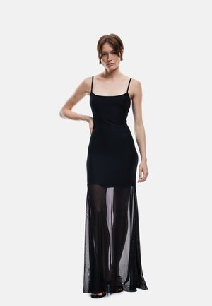 Mujer de pie con vestido negro sin mangas con una superposición transparente hasta el suelo y tacones abiertos negros sobre un fondo blanco.