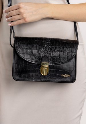 Frau hält schwarze Schultertasche aus Leder mit Krokodillederstruktur und goldenem Verschluss, trägt ein beiges Kleid, Nahaufnahme von Oberkörper und Händen.