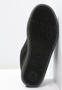 Schwarze Skate-Schuhsohle mit strukturiertem Gummioberfläche, geometrischen Mustern und einem markierten Emblem mit Grip-Details für verbesserte Traktion.
