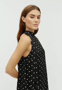 Rochie neagră fără mâneci cu buline albe. Are un guler înalt și un design drapat, fiind realizată dintr-un material neted și ușor.