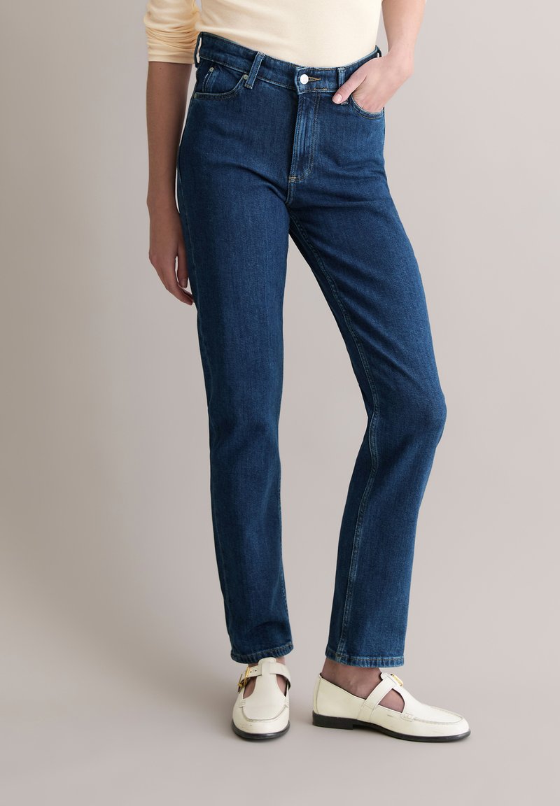 Jean droit en denim bleu foncé avec taille haute, design à cinq poches, passants pour ceinture et braguette classique à glissière. Associé à des chaussures blanches.