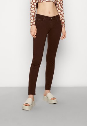 Kvinde iført mørkebrune skinny jeans med høj talje, cropped langærmet top med tusindfryd-mønster og off-white platform-sandaler, stående på gråt gulv.