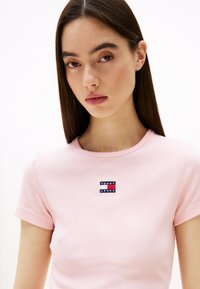 Ružová rebrovaná tričko s okrúhlym výstrihom a krátkymi rukávmi, s modrým a červeným obdĺžnikovým logom, na ktorom je nápis "TOMMY JEANS."