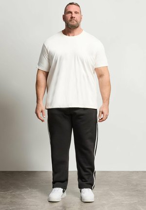 Hombre con barba de pie, vistiendo una camiseta blanca lisa, pantalones de chándal negros con rayas blancas a los lados y zapatillas blancas sobre un fondo claro.