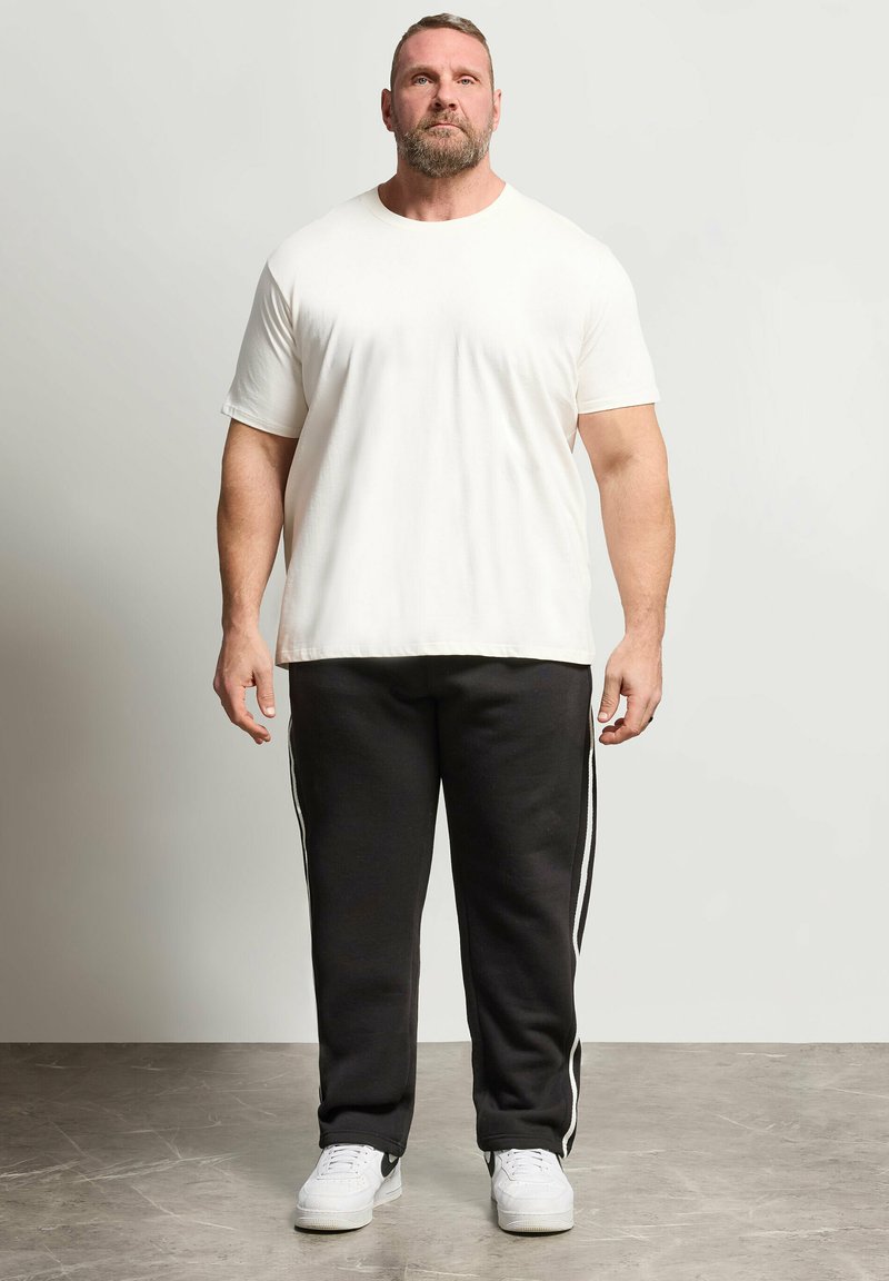 Uomo barbuto in piedi, che indossa una maglietta bianca semplice, pantaloni tuta neri con strisce bianche laterali e scarpe da ginnastica bianche su uno sfondo chiaro.
