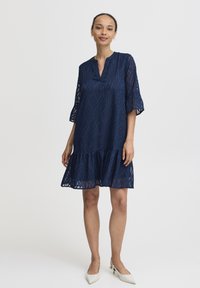 Rochie de culoare bleumarin cu un material brodat cu un model de frunze, mâneci trei sferturi și tiv ondulat. Prezintă o croială lejeră și decolteu în V.