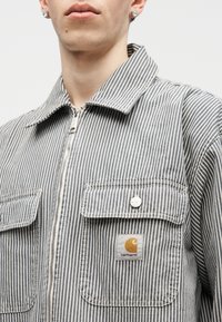 Mees, kes kannab halli ja valget vertikaalset triibulist lukuga jope, millel on kaks rinnataskut, üks neist Carhartti logo plaastriga.