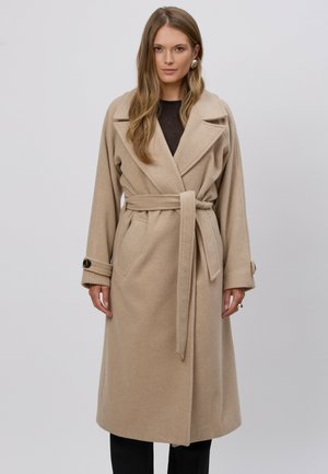 Kobieta o długich włosach, ubrana w beżowy wełniany płaszcz z paskiem i czarną bluzkę, stojąca na tle jednolitego jasnoszarego tła.