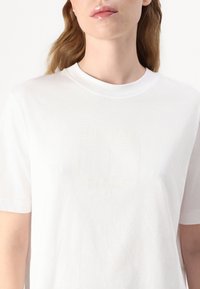 Bijela pamučna majica s okruglim izrezom, s diskretnim utisnutim logotipom i natpisom "HILFIGER NEW YORK 1985" ispod.