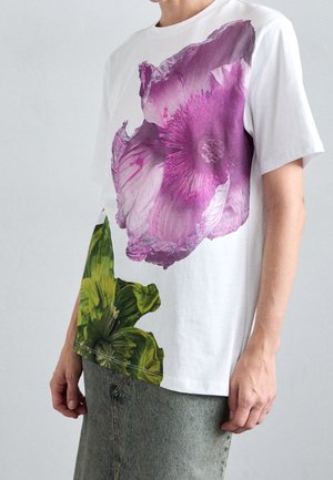 Weißes kurzärmeliges Hemd mit großen lila und grünen Blumenmustern, getragen von einer Person mit heller Haut und blau-grauem Jeansrock.