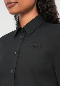 Melna pogām aizdares krekls no gludas auduma ar smailu apkakli un izšūtu logotipu kreisajā krūšu daļā, melnas pogas.