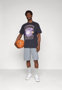 Čierne grafické tričko s žltým a fialovým textom "NIKE LOOPS", v kombinácii so svetlosivými šortkami a bielymi teniskami, držiacimi basketbal.