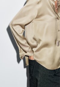 Beige satin long-sleeve blouse tucked slightly into dark denim pants, with hand in pocket and layered silver necklaces.