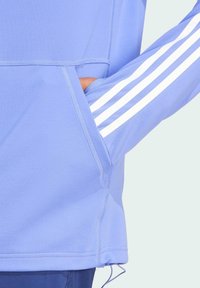 Hellblauer Sportkapuzenpullover mit Kängurutasche. Verfügt über weiße Streifen an den Ärmeln und einen strukturierten Stoff. Verstellbarer Saum mit Kordelzug.