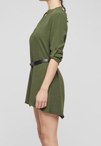 Femme en vue de profil portant une robe verte à manches longues avec les manches retroussées et une ceinture noire à la taille sur un fond uni.