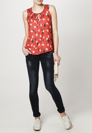 Blouse sans manches rouge avec un col froncé et des motifs abstraits blancs et bleus, associée à un jean skinny noir et des chaussures blanches.