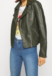 Olivgrön läderbikerjacka med silverfärgade dragkedjor, flera fickor och vadderade detaljer på axlarna; över en grafisk t-shirt.