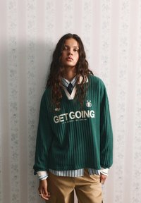 Zelený pruhovaný oversized sveter s textom "GET GOING", navrstvený cez biele a zelené pruhované tričko, spárovaný s béžovými nohavicami.