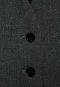 Grau maßgeschneiderte Blazer aus strukturiertem Stoff, mit zwei schwarzen Knöpfen und einem Reverskragen. Nahaufnahme fokussiert auf die Knopfdetails.