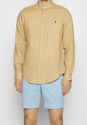 Man die een beige overhemd met lange mouwen en knopen draagt, met een klein logo op de borst en lichtblauwe korte broek, staand tegen een effen achtergrond.