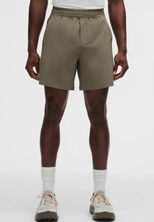 Man met olijfgroene shorts met elastische taille, witte sokken tot halverwege de kuit en beige hardloopschoenen, staand tegen een effen achtergrond.