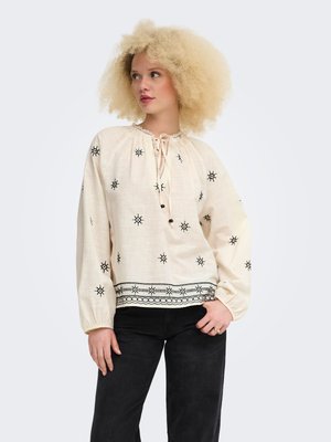 Femme aux cheveux blonds bouclés portant une blouse crème avec des motifs brodés noirs et un pantalon noir, posant avec une main dans la poche.
