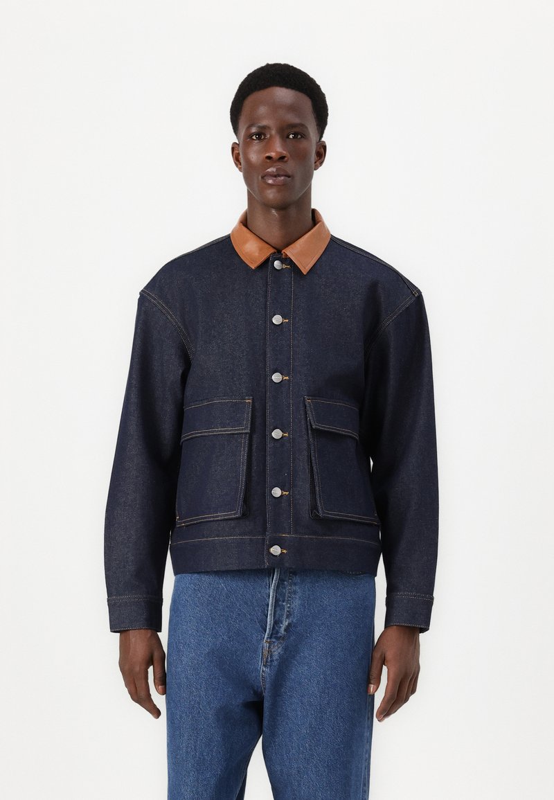 Veste en denim avec un corps bleu foncé, un col en cuir marron, des boutons-pression et deux grandes poches avant, avec des détails de couture visibles.