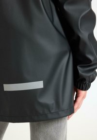 Giacca nera impermeabile con polsini elasticizzati, dotata di una striscia riflettente argentea vicino all'orlo; texture liscia e vestibilità comoda.