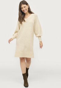 Beige gebreide jurk met v-hals, ballonmouwen, geribbelde manchetten en zoom. Kniehoogte met een zachte, gestructureerde uitstraling.