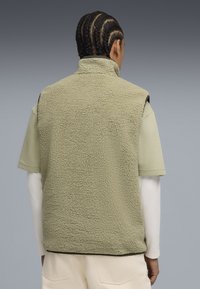 Gilet in pile verde con una superficie testurizzata, colletto alto e design senza maniche, indossato sopra una camicia a maniche corte verde chiaro.