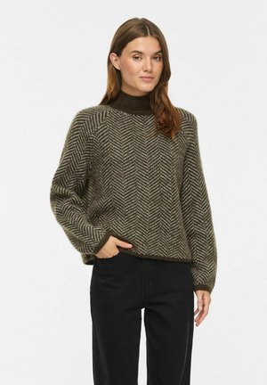 Femme portant un pull tricoté marron à motif chevrons avec manches longues et un pantalon noir, debout avec une main dans la poche.