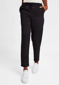Pantalon noir léger avec une taille à cordon, présentant un motif de petits pois rouges et blancs ; porté avec des baskets blanches.