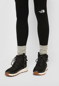 Black insulated boots with white soles and textured laces, paired with black leggings featuring a logo and gray knitted cuffs.