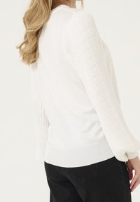 Vit tröja med ribbad textur på ärmarna och slät kropp. Har en rundad halsringning och åtsittande nedre del, tillsammans med svarta byxor.