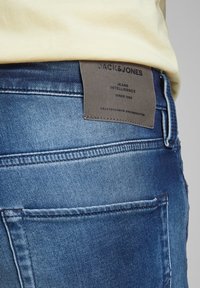 Primer plano del bolsillo trasero de unos jeans de mezclilla azul y la etiqueta de la marca Jack & Jones en la cintura, debajo de una camisa amarilla.
