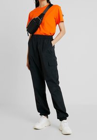 Pantalones cargo negros con puños elásticos, combinados con una camiseta de color naranja brillante. Se lleva un bolso negro tipo bandolera al hombro. Zapatillas blancas completan el conjunto.