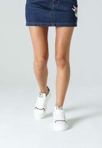 Vita sneakers med burgundiska accenter och logodetalj. Synliga sömmar och texturerat material. Bärs med en jeanskjol med broderi.