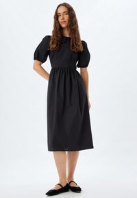 Robe noire, longueur genou, avec des manches bouffantes, un col, et une taille froncée. Tissu lisse, corsage ajusté, et jupe évasée.