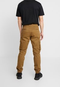 Bruine cargobroek met taps toelopende enkels, zijzakken en een gladde textuur. Draagt een eenvoudige zwarte t-shirt en sneakers.