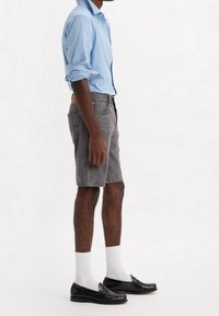 Hombre con una camisa de manga larga azul claro arremangada, pantalones cortos grises hasta la rodilla, calcetines blancos y mocasines de cuero negros de pie de lado.