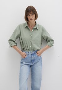 Helles grünes Hemd mit dezentem Pünktchenmuster, kombiniert mit hoch taillierten, hellblauen Jeans; umgeschlagene Ärmel.