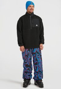 Zwarte fleece pullover met ritsenkraag en voorkeurszak, gecombineerd met blauwe en roze gestreepte sneeuwbroeken. Zwarte skibotten maken de look compleet.