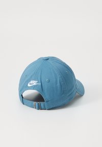 Sininen puuvillainen baseball-lippis säädettävällä hihnalla, jossa on valkoinen Nike-logo brodeerattu taakse ja pyöreä kruunu.