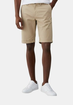 Uomo che indossa pantaloncini beige fino al ginocchio e sneakers bianche, in piedi con una mano in tasca davanti a uno sfondo semplice.
