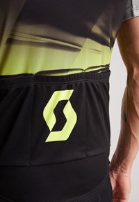 Černý sportovní dres s gradientním žlutým designem, vyrobený z texturované tkaniny, s výrazným žlutým logem na zadní kapsě.