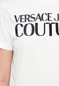 Balta medvilninė marškinėliai su ryškiu juodu iškilniu tekstu "VERSACE JEAN COUTURE" per krūtinę, su apvalia apykakle ir trumpomis rankovėmis.