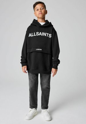 Chlapec stojící v černé mikině "ALLSAINTS LONDON", tmavě šedých džínách a bílých teniskách na jednobarevném šedém pozadí.