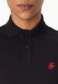 Juodas polo marškinėlis su tekstūruotu audiniu, trumpu apykakle ir dviem mygtukais, su raudonu siuvinėtų logotipu ant krūtinės.