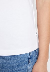 T-shirt en coton blanc avec un col rond, des manches courtes et une petite étiquette de marque sur la couture latérale gauche. Porté avec un short en denim bleu.