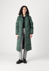 Lang, grønn pufferjakke med glidelås og hette, har et quiltet design. Brukt over en svart topp og grå jeans, parret med svarte sko.
